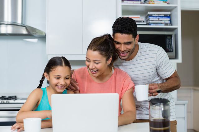 Family enjoying time together in kitchen, using laptop and having coffee. Ideal for promoting family bonding, technology use in daily life, and home lifestyle. Suitable for advertisements, blogs, and articles about family life, technology, and home activities.