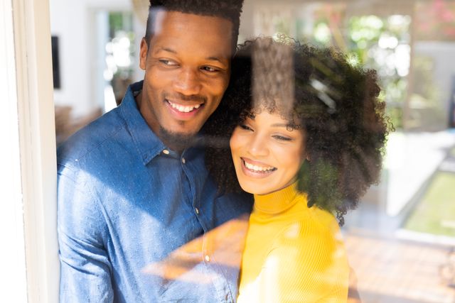 This image depicts a happy biracial couple embracing and smiling while looking out of a window at home. It conveys themes of love, togetherness, and inclusivity. Ideal for use in advertisements, blogs, or articles related to relationships, domestic life, and leisure time.