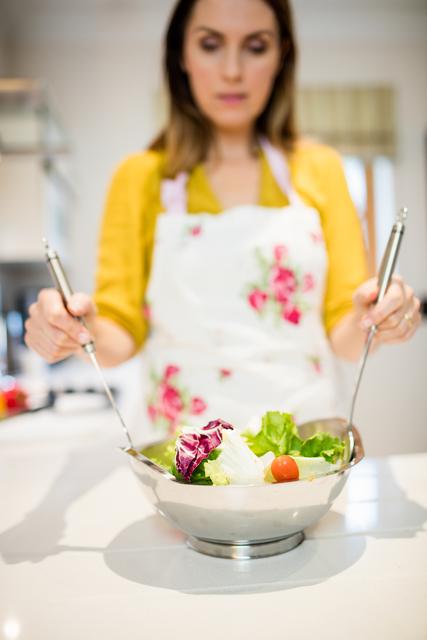 Woman preparing a fresh salad in a modern kitchen at home. She is wearing a floral apron and using salad tongs to mix the ingredients. This image can be used for promoting healthy eating, cooking blogs, lifestyle articles, and domestic life themes.
