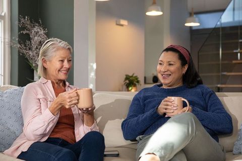Two senior women enjoying coffee and conversation sharing warm mugs on cozy living room sofa at home