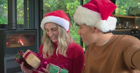 Couple exchanging christmas gifts wearing santa hats and sweaters by wood-burning stove
