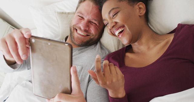 Couple enjoying time together while watching something on a tablet in bed. Ideal for topics relating to relationships, technology use in leisure time, or mixed-race couples. Can be used in advertisements for tech gadgets, relationship advice blog posts, or relaxation products.