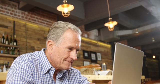 Senior man sitting at cafe using laptop, dressed in a checked shirt. Rustic cafe atmosphere with wooden walls and industrial light fixtures. Suitable for themes like technology use among seniors, remote working, casual dining, and mature lifestyle.