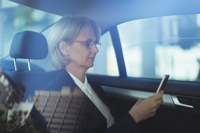 Senior businesswoman in formal attire using smartphone while traveling in car. Ideal for illustrating corporate travel, business communication, executive lifestyle, and professional work environments.