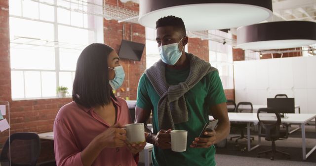 Two colleagues wearing face masks are holding coffee mugs and engaging in a discussion in a modern office with large windows and contemporary design elements. This image can be used to represent workplace safety, teamwork, and professional environments adhering to health guidelines.