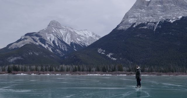 Solitary person ice skating on a frozen lake surrounded by tall, snow-capped mountains and forests. Captures winter serenity and the quiet beauty of nature. Ideal for use in outdoor adventure promotions, winter sports advertising, and travel brochures emphasizing peaceful, scenic destinations.