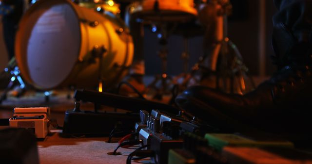 Close-up of a musician's foot in a dark music studio, adjusting effect pedals on the floor near a drum set. This image is ideal for use in promotions for music studios, live performances, band practices, and music equipment advertisements.