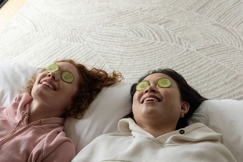 Two friends smiling and relaxing with cucumber slices on eyes, hoodies, cozy pampering