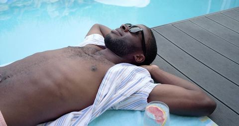 Relaxing shirtless man sunbathing by pool with sunglasses, striped shirt and citrus drink