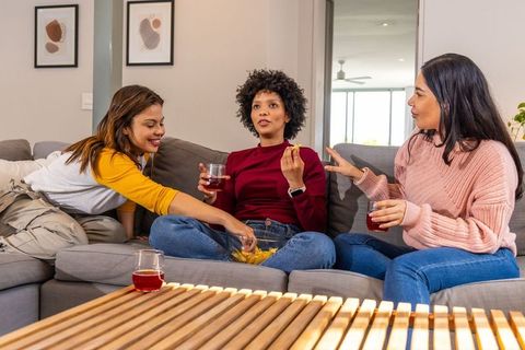 Diverse friends sitting on sofa sharing snacks and red drinks in cozy homey living room