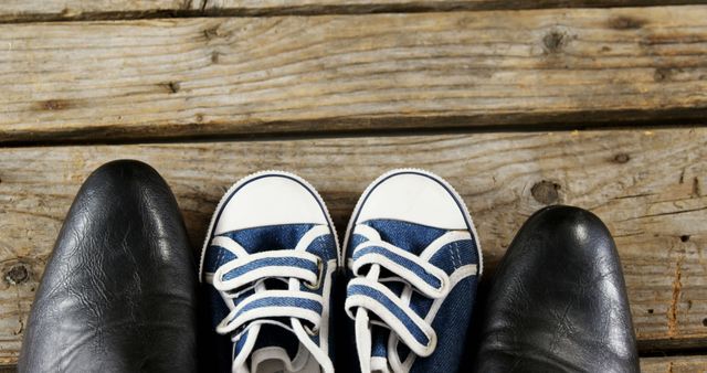 A pair of adult black leather shoes stands next to a child's blue and white sneakers on a wooden floor, with copy space. It symbolizes the concept of childhood and growth or the bond between parent and child.