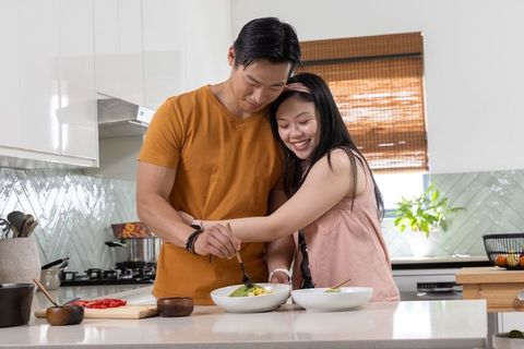 Asian couple cooking together on bright modern kitchen island sharing healthy salad moment