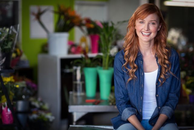 Happy female florist with red hair sitting on table in flower shop, smiling. Ideal for use in articles or advertisements about small businesses, entrepreneurship, floral arrangements, and retail environments. Perfect for promoting flower shops, business success stories, and cheerful work environments.