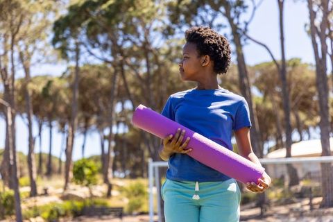 African american youth holding purple yoga mat in sunlit park in blue tee, teal shorts
