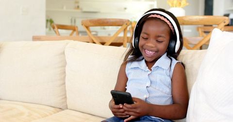 Joyful Girl Sitting on Couch Wearing Headphones Engaged with Smartphone