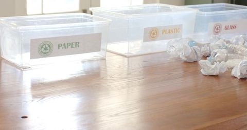 Clear recycling bins labeled paper plastic glass on sunlit wooden table