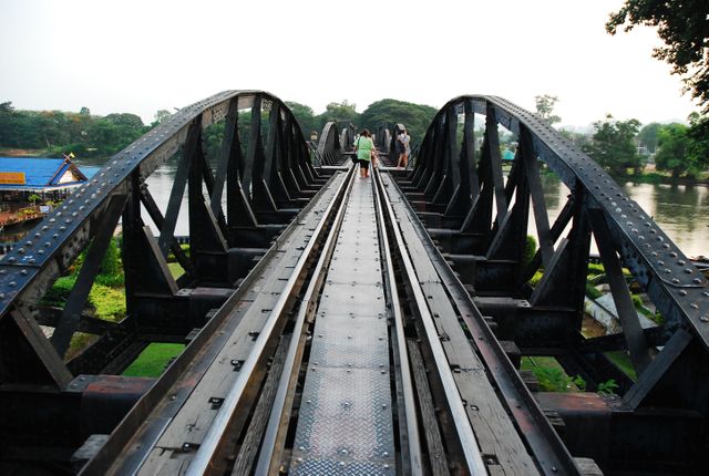 Tourists are walking on a historic steel railway bridge spanning over a river. The bridge is constructed with large iron beams and displays a classic industrial design. This image can be used for topics related to historical landmarks, travel destinations, architectural engineering, and outdoor activities.