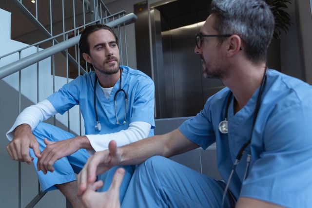 Two male surgeons in blue scrubs are engaged in a conversation while sitting on hospital stairs. Both wearing stethoscopes, they seem to be discussing important medical matters or taking a short break from their duties. This image can be used for illustrating teamwork and collaboration among healthcare professionals, medical discussions, or hospital environments.
