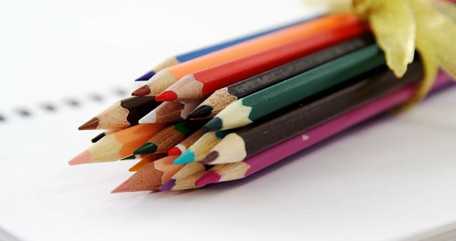 A bundle of sharpened colored pencils is tied together with a golden ribbon, resting on a blank notebook, with copy space. Colored pencils are essential tools for artists, students, and anyone engaged in creative work.
