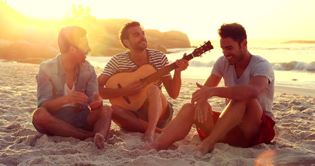 Three young men sit on sandy beach at sunset, one playing guitar while others smile and enjoy relaxing conversation. Perfect for themes of friendship, summer fun, leisure activities, and outdoor relaxation.