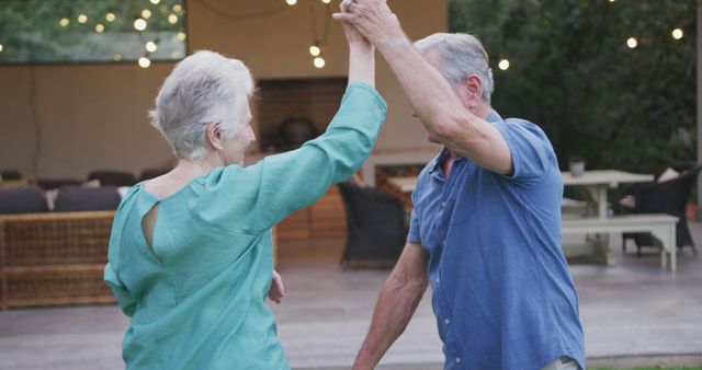An elderly couple is seen dancing joyfully outdoors, with string lights illuminating the evening. Perfect for themes showcasing love, happiness, and active senior living. Suitable for advertisements, lifestyle articles, and promotional materials celebrating romance and enjoyment among retirees.