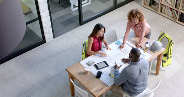 Group of diverse architects and engineers collaborating on a building project in a modern office setting. They are discussing construction plans and blueprints spread on a table, using digital tablets and taking notes. Ideal for use in articles or advertisements about teamwork, architecture, engineering projects, office environments, professional collaboration, and diversity in the workplace.