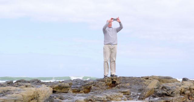 Man standing on rocky beach taking photos with smartphone. Sky filled with light clouds in bright day. Ideal for travel blogs, advertisements for outdoor activities, nature photography, promotional material for tourist destinations or beach resorts.