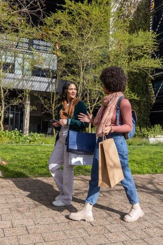 Two friends walking and chatting on sunny urban campus carrying shopping bags, backpacks