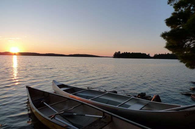 Two canoes rest beside a tranquil lake during sunset. The peaceful scene features vibrant colors of the setting sun reflecting on water. Ideal for promoting outdoor activities, adventure sports, vacation getaways, travel destinations, and nature-related themes.