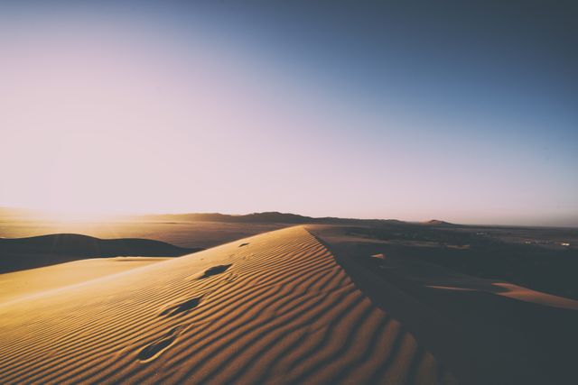 Footprints on sand dunes leading into the horizon under a captivating sunrise in a serene desert setting. Ideal for travel blogs, adventure advertisements, nature documentary visuals, or inspirational and motivational material relating to journeys and exploration.