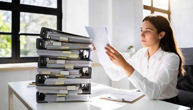 Office worker reviewing large stack of binders and documents at sunlit desk