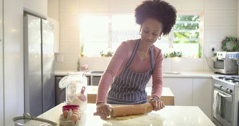 African American woman rolling dough on countertop in sunlit modern kitchen, striped apron