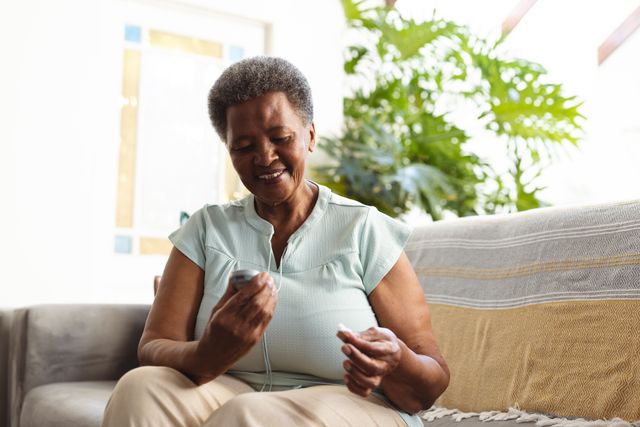 Senior African American woman sitting comfortably on sofa, smiling while using glucometer to check blood sugar. Good for promoting diabetes management, elderly health care, medical equipment for seniors, and healthy living in retirement.