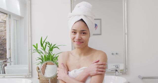 Woman wrapping towel around her body in bathroom, smiling towards camera. Mirror and plant in background create a natural, clean atmosphere. Ideal for use in self-care, personal hygiene, and skincare advertisements.