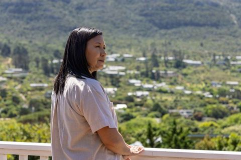 Mature asian woman standing on balcony gazing over sunlit valley rooftops and hillside
