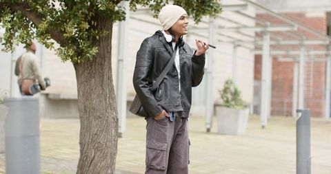 Young man speaking into smartphone on campus wearing beanie headphones leather jacket