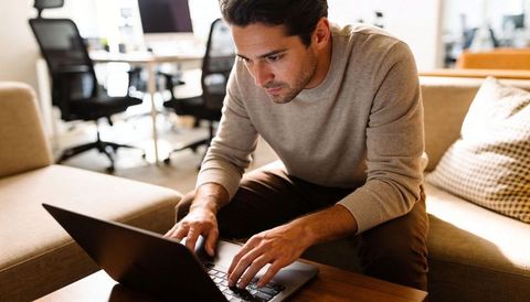 Focused young man typing on laptop in modern coworking lounge, casual sweater, daylight