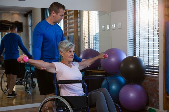Senior woman in wheelchair exercising with dumbbells under the guidance of a physiotherapist in a clinic. Ideal for use in healthcare, rehabilitation, physical therapy, and senior fitness content.