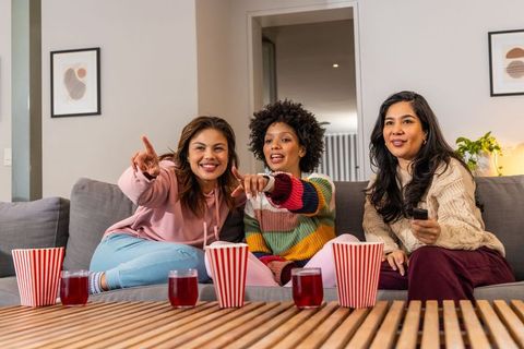 Multicultural women enjoying movie night on cozy couch, pointing at screen and laughing