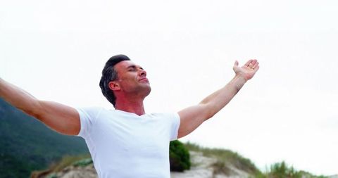 Midlife man stretching arms wide, breathing deeply on coastal dune, embracing freedom