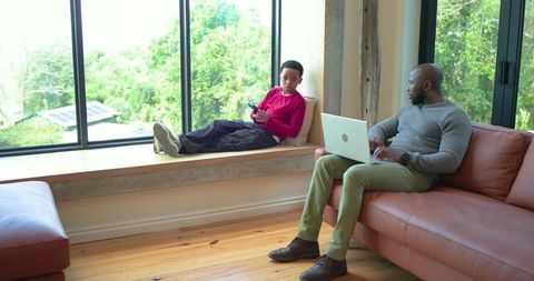 African american father and son relaxing on window bench and sofa, using laptop and smartphone