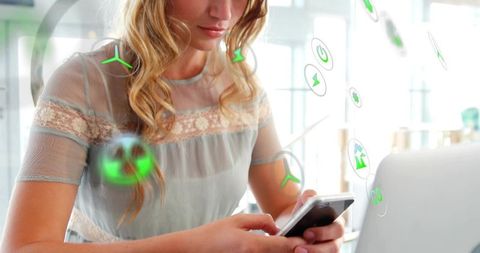 Young woman checking smartphone at desk with laptop and floating green eco app icons