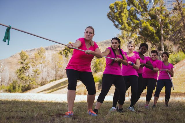 Group of women participating in a tug of war competition during an outdoor boot camp training session. They are wearing matching pink shirts and are demonstrating teamwork and strength. This image can be used for promoting fitness programs, team-building activities, or outdoor sports events.