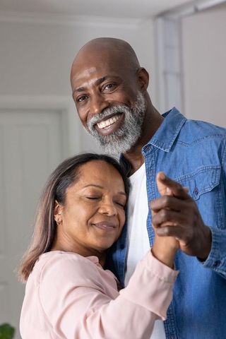 Smiling mature couple holding hands and slow dancing in cozy sunlit living room