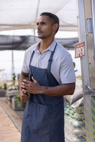 Nursery owner standing at greenhouse entrance wearing denim apron and holding tablet