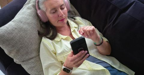 Mature woman reclining on sofa listening with pink headphones and scrolling smartphone