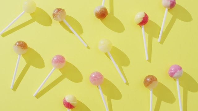 Bright multicolored lollipops arranged on vibrant yellow background, casting playful shadows. Suitable for use in advertisements, packaging designs, confectionery promotions, children's party invitations, or digital wallpapers emphasizing fun and vibrancy.