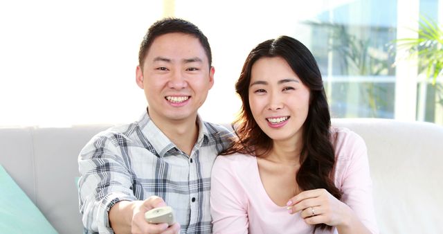 A young Asian couple smiles at the camera while sitting on a couch, enjoying a relaxed day at home. Their cheerful expressions suggest a moment of leisure or shared entertainment, with a remote control in hand indicating they might be watching television.