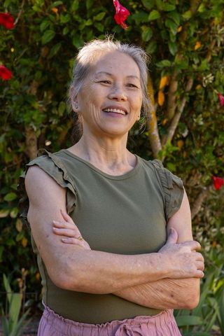 Senior asian woman standing in sunlit garden smiling with arms crossed by hibiscus hedge