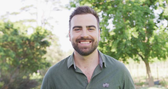 Young man standing outdoors in casual shirt smiling at camera with lush green trees in background. Suitable for concepts related to happiness, lifestyle, relaxation, youth, and outdoor activities.